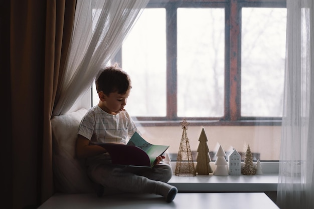 Chico lindo está leyendo un libro sentado en el alféizar de la ventana cerca de la ventana Casa acogedora con árboles de Navidad decorativos Dulce hogar Estilo de vida de vacaciones de invierno