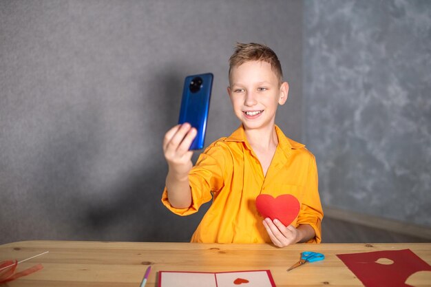 Un chico lindo está sentado en una mesa y haciendo tarjetas de San Valentín Tomando selfies