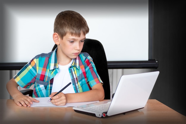 Un chico lindo está sentado en una mesa, mirando una computadora portátil, escribiendo tareas o preparándose para un examen. Concepto Regreso a la escuela. Copia espacio
