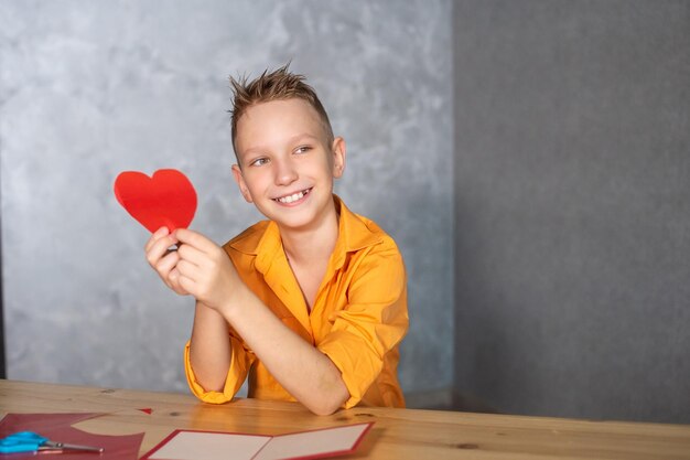 Un chico lindo está sentado en una mesa y sostiene un corazón rojo en sus manos para una tarjeta de San Valentín