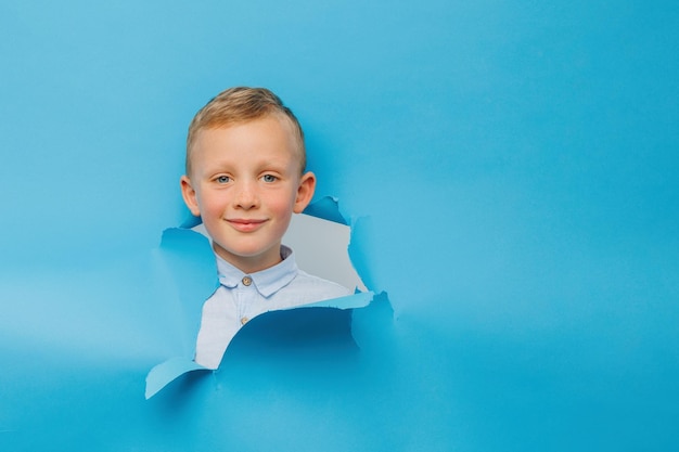 El chico lindo feliz se divierte jugando en la pared de fondo azul sube a través de un agujero en el papel