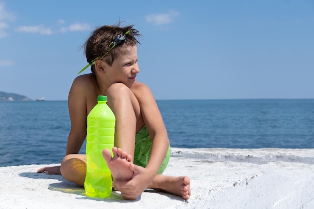 Un chico lindo con gafas de natación y pantalones cortos con una botella de agua se sienta en un muelle a la orilla del mar.
