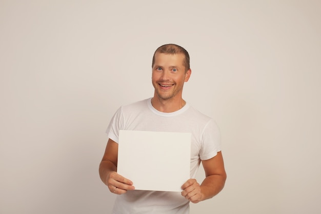 Un chico lindo joven con una camiseta blanca sostiene una hoja de papel blanco en sus manos
