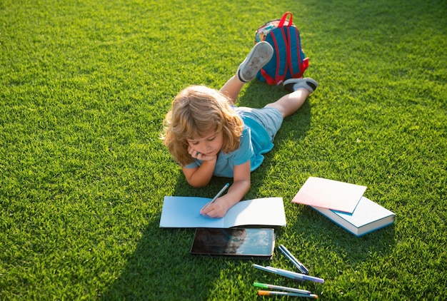 Chico lindo leyendo un libro tendido en la hierba escribiendo notas en el cuaderno niño leyendo un libro en el verano