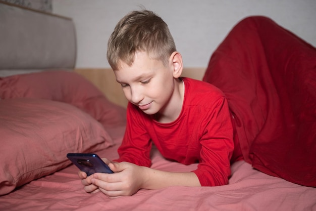 Un chico lindo en pijama rojo está acostado en la cama debajo de una manta y jugando por teléfono