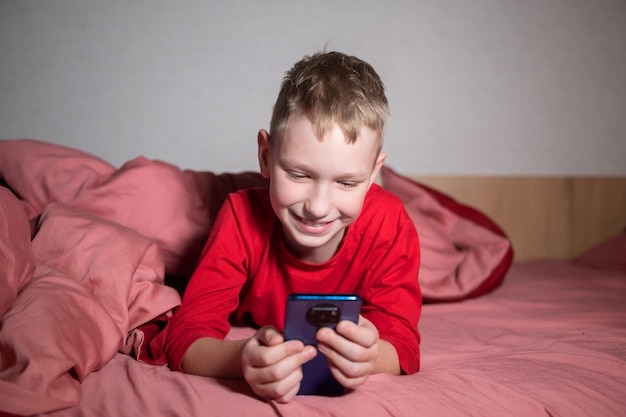 Un chico lindo en pijama rojo está acostado en la cama debajo de una manta y mirando el teléfono.