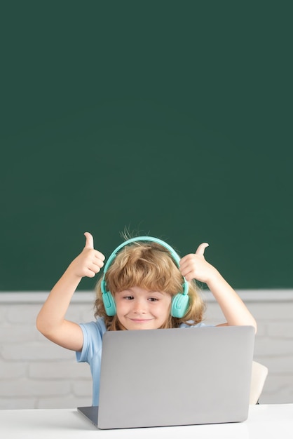 Un chico lindo con los pulgares hacia arriba usando audífonos estudia con una laptop en el aula escuchando el curso de lecciones de audio Primer día en la escuela