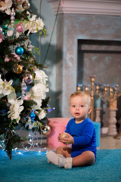 Chico lindo sentado en la alfombra cerca del árbol de Navidad