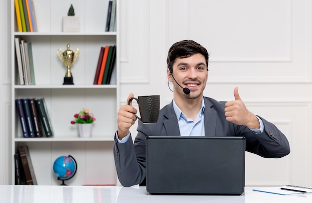 Chico lindo de servicio al cliente en traje gris con computadora y auriculares sosteniendo una taza y sonriendo