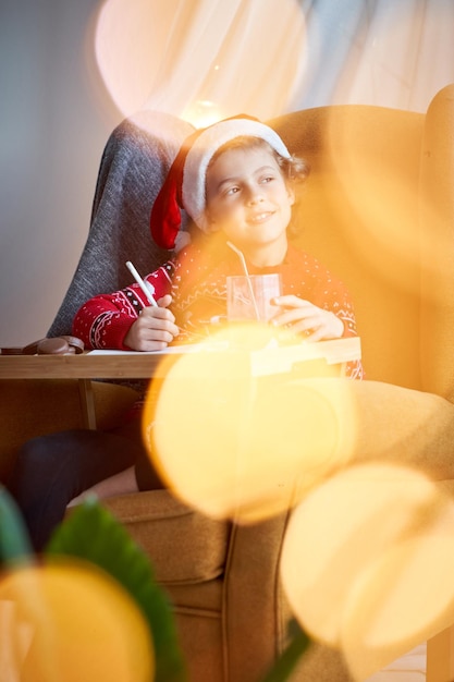 Un chico lindo con sombrero de Papá Noel mirando a lo lejos mientras escribe una carta a Papá Noel en una rom ligera con bokeh durante la celebración de Navidad