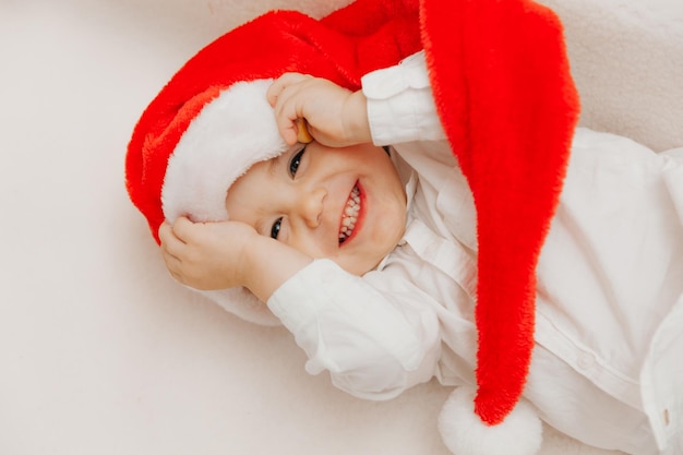 Chico lindo con sombrero de santa claus se encuentra en una manta cerca del árbol de Navidad. niño feliz se encuentra cerca del árbol de Navidad y sonríe
