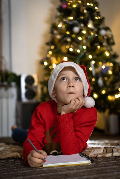 Chico lindo en suéter rojo y gorro rojo de santa escribe una carta a Santa y pensando en regalos