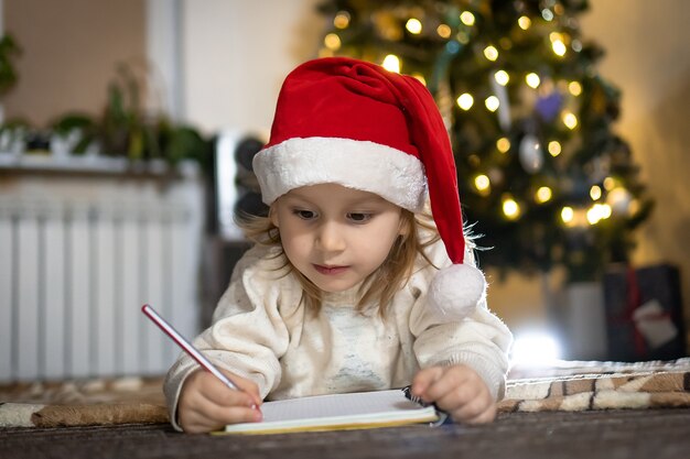 Chico lindo en suéter rojo y sombrero rojo de santa escribe una carta a Santa