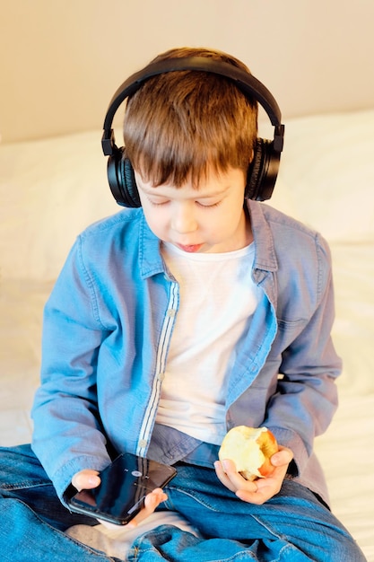 Chico lindo con teléfono y auriculares escuchando música en el interior sentado en la cama en el dormitorio Niño come una manzana