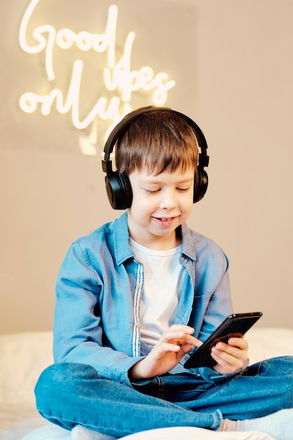 Chico lindo con teléfono y auriculares escuchando música en el interior sentado en la cama en el dormitorio