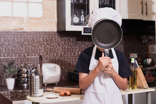 Un chico lindo con uniforme de chef que cubre la cara con una sartén de teflón en la cocina. Concepto de comida y cocina.