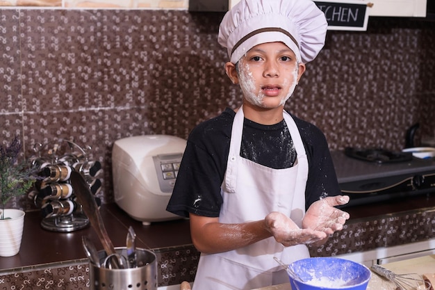 chico lindo usando uniforme de chef mostrando harina en sus manos y mejillas mientras hace masa en la cocina