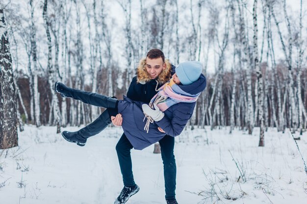 Chico llevando a su novia en las manos en el bosque de invierno. Gente divirtiéndose al aire libre