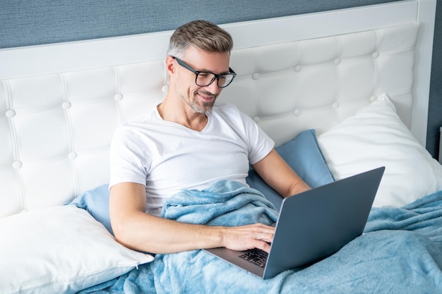 Foto chico maduro con gafas trabajando en una laptop en la cama