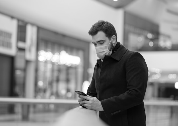 Chico con mascarilla y abrigo está usando un teléfono móvil en el centro comercial de la ciudad en tiempo de pandemia