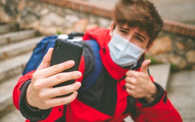 Chico con mascarilla y mochila tomando un selfie en la calle en un día de invierno