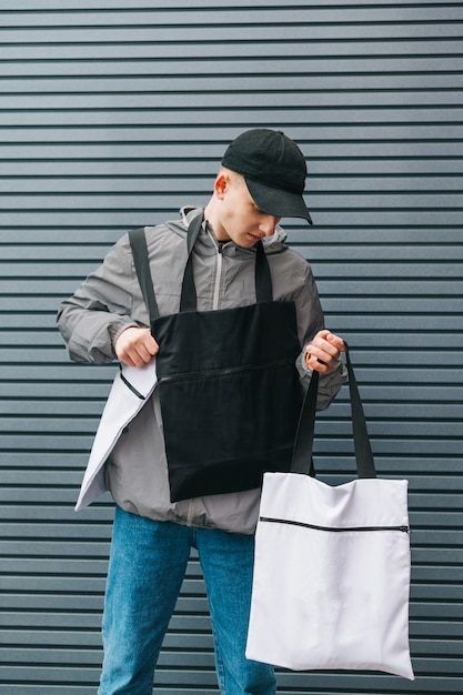 Chico de moda en ropa elegante posando con bolsas ecológicas en sus manos en un gris