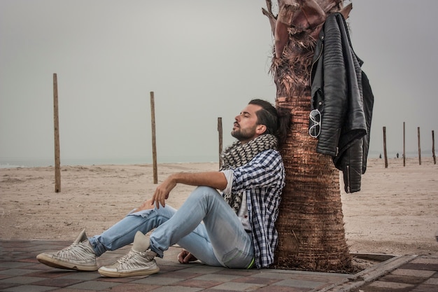 Foto chico de moda sentado junto al mar cerca de una palma