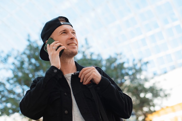 Chico moderno de moda joven, un hombre con una gorra negra y gafas de sol con un teléfono inteligente, habla por teléfono sobre un fondo urbano de la ciudad en un túnel en los rayos del sol al atardecer.