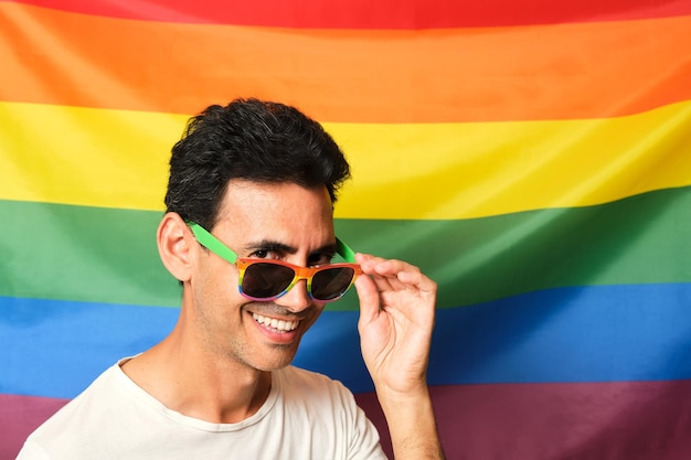 Chico moreno sonriente con camiseta blanca con gafas de arco iris lgbtq mirando por encima de las gafas