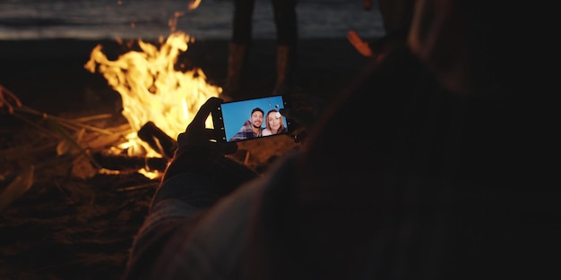 Chico muestra a chica una foto en su teléfono junto a una fogata en la playa