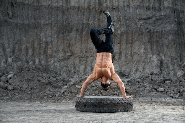 Chico musculoso con el pecho desnudo en equilibrio mientras está de pie con las manos en el neumático grande. Hombre activo con máscara negra y pantalones deportivos. Concepto de resistencia y fuerza.