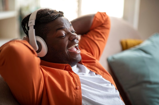 Chico negro alegre recostado en el sofá cantando canciones con auriculares