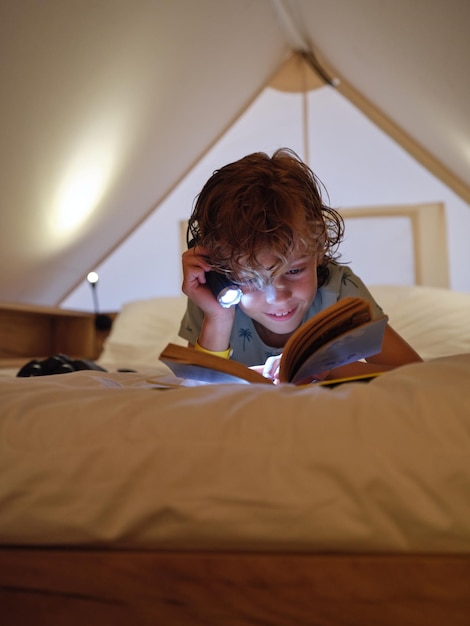 Un chico optimista con una linterna brillante leyendo un libro fascinado mientras yacía en una cama blanca en una mansarda en casa por la noche