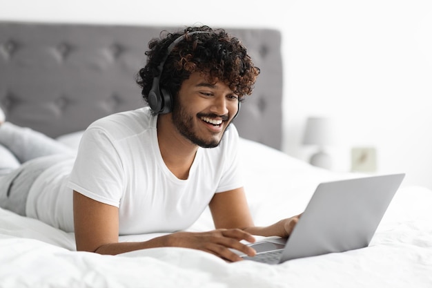 Chico oriental relajado disfrutando el fin de semana en la cama usando una computadora portátil