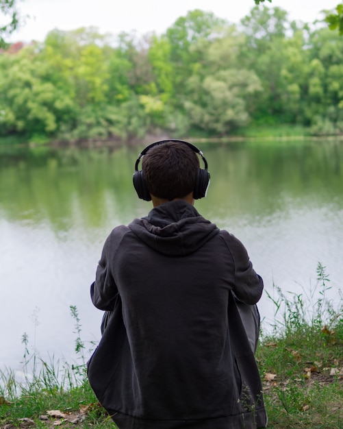 El chico en la orilla del río escuchando música. Lejos de la ciudad circundante. Escuchar música auriculares