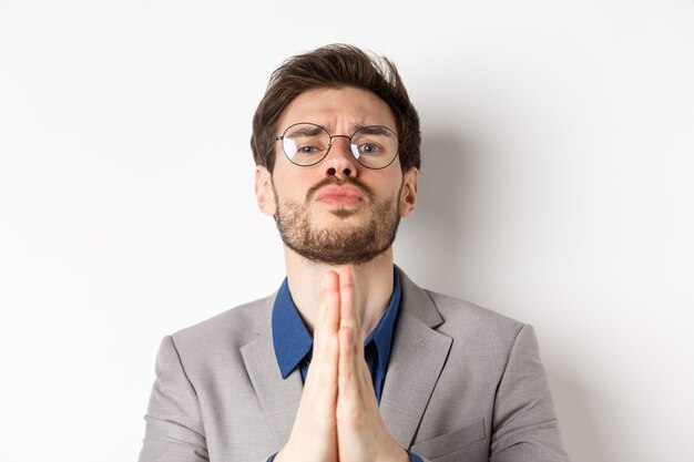 Chico pegajoso con gafas pidiendo el último favor, pidiendo ayuda y mirando tristes ojos lindos, de pie sobre fondo blanco.