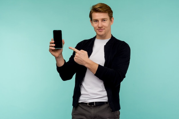 Chico pelirrojo deportivo alegre muestra un dedo en la pantalla del teléfono inteligente sobre un fondo azul. - imagen