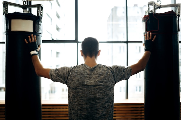 Chico de pelo oscuro se encuentra entre dos sacos de boxeo contra el fondo de ventanas panorámicas en el gimnasio de boxeo.