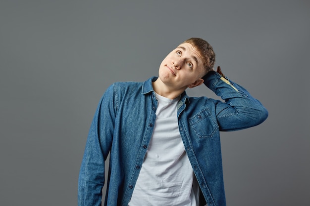 Chico pensativo vestido con camiseta blanca y una camisa de jeans tiene su mano sobre su cabeza sobre el fondo gris en el estudio.