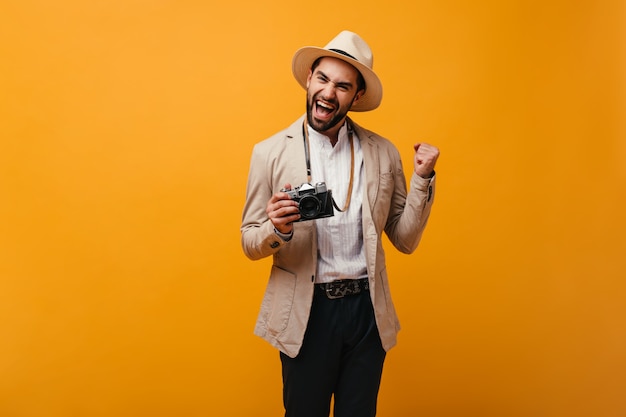 Chico positivo posando felizmente en la pared naranja y sosteniendo una cámara retro