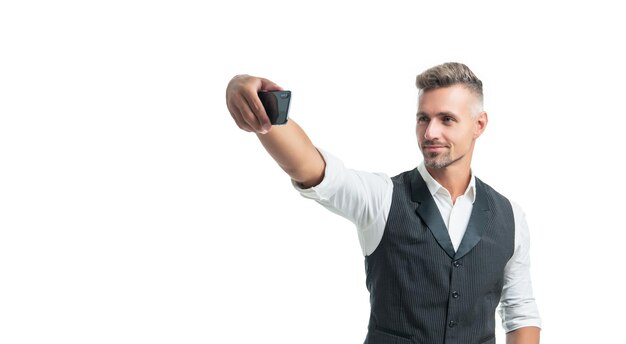 Chico profesional feliz en chaleco de traje tomando selfie con teléfono inteligente aislado en blanco