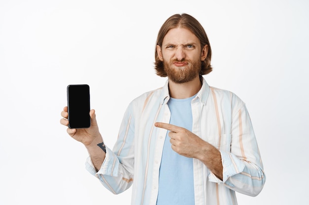 Chico reacio con cabello rubio, señalando con el dedo a la pantalla del teléfono móvil y haciendo muecas, no le gusta la aplicación, mala interfaz, de pie contra el fondo blanco