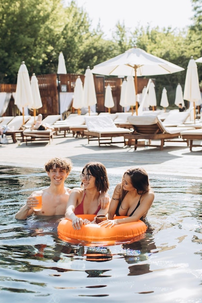 Chico rizado y dos hermosas chicas bebiendo cócteles y divirtiéndose junto a la piscina en verano