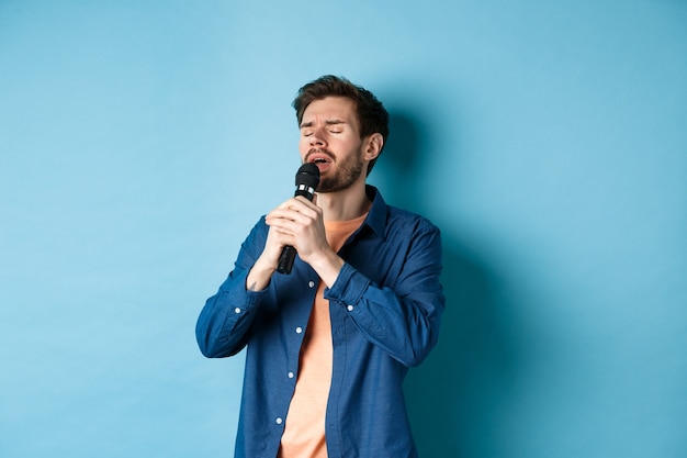 Chico romántico cantando en el micrófono, actuando en el escenario, de pie sobre fondo azul.