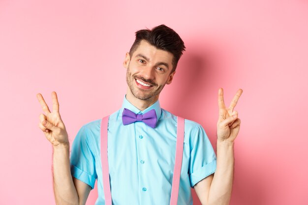 Chico romántico con pajarita y tirantes mostrando signos de paz y sonriendo a la cámara, vestido con ropa para la fecha, de pie sobre fondo rosa.