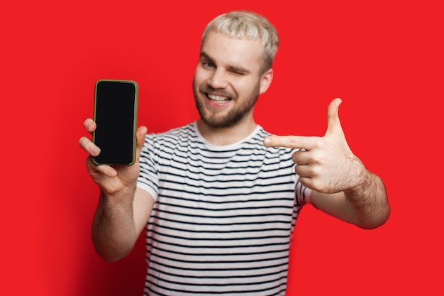 Chico rubio caucásico con barba corta apuntando al teléfono con espacio en blanco sobre fondo rojo.