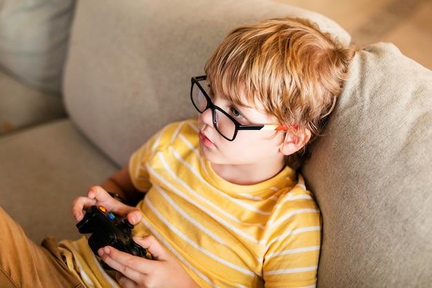 Chico rubio con gafas grandes jugando en la consola sentado en el sofá. Niño europeo que tiene un hobby