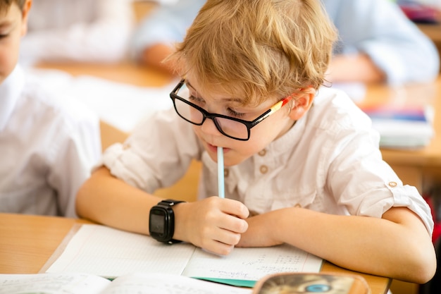 Chico rubio con grandes gafas negras sentado en el aula, estudiando, sonriendo. Educación en la escuela primaria, primer día en la escuela.