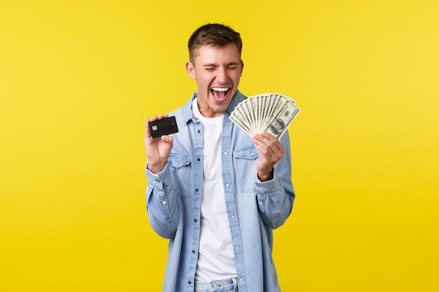 Chico rubio guapo feliz emocionado mostrando dinero y tarjeta de crédito, gritando sí regocijo, celebrando logros, recibió dinero extra, ganando lotería, fondo amarillo de pie.