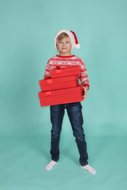 Un chico rubio guapo con un suéter de Navidad, sombrero de Santa Claus tiene cajas de regalo en sus manos